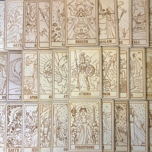 Op de afbeelding: Een verzameling houten tarotkaarten met ingewikkelde gegraveerde illustraties. De kaarten tonen afbeeldingen van goden en symbolen, waaronder Athena, Brigid en Persephone. De kaarten zijn in een verspreid patroon gerangschikt, waarbij de gedetailleerde kunst en de natuurlijke houtnerf worden getoond.