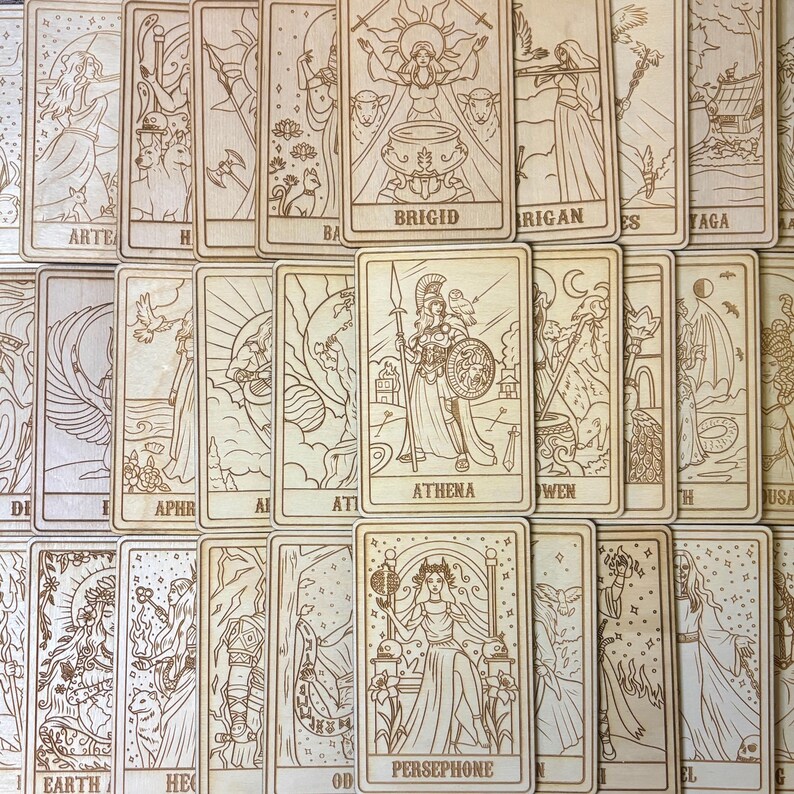 Op de afbeelding: Een verzameling houten tarotkaarten met ingewikkelde gegraveerde illustraties. De kaarten tonen afbeeldingen van goden en symbolen, waaronder Athena, Brigid en Persephone. De kaarten zijn in een verspreid patroon gerangschikt, waarbij de gedetailleerde kunst en de natuurlijke houtnerf worden getoond.