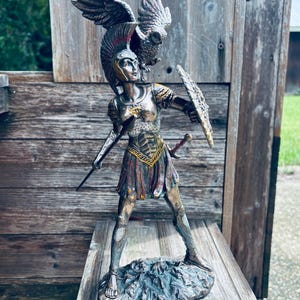 May include: A bronze-toned statue of a warrior figure, adorned with a helmet and shield, and an owl perched on the warrior's shoulder. The warrior holds a sword and spear, standing on a textured base. The statue has intricate details and a weathered appearance.
