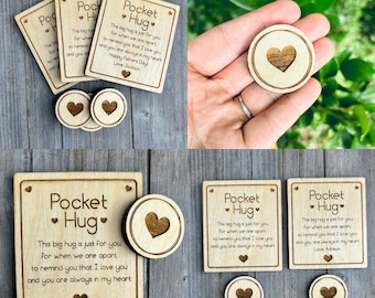 May include: Wooden Pocket Hug tokens and cards. The cards have the text "Pocket Hug" and a heart design. The text on the cards reads "This big hug is just for you..." and includes a name. The tokens have a heart design.
