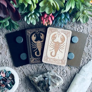 May include: Two rectangular magnets with a zodiac theme. One is black with a gold scorpion design, and the other is light brown with a scorpion design. The word "SCORPIO" is at the bottom of the light brown magnet.