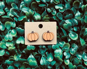 May include: A pair of orange pumpkin-shaped earrings made of wood, displayed on a tan card. The earrings have a simple, carved design and are set against a backdrop of green foliage.