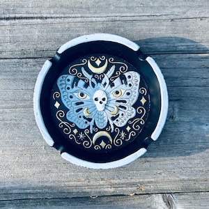 May include: Round black ashtray with a white rim. The ashtray features a detailed moth design with a skull, moons, and stars in shades of blue, gold, and white. The ashtray is made of a durable material and is suitable for indoor or outdoor use.