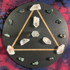 May include: A black circular wooden board with a gold triangle design. The board is decorated with clear quartz crystals and green gemstones.