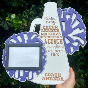 May include: White wooden megaphone with a purple pom-pom on each side. The megaphone has a picture frame in the center and the text "Behind every cheer leader who believes in themselves is a coach who believed in them first. Coach Amanda".