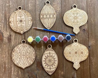 May include: Six unfinished wooden ornaments in the shape of Christmas tree ornaments with intricate designs. The ornaments are arranged in two rows with a row of six paint pots in between. The paint pots are yellow, green, red, blue, brown, and black.