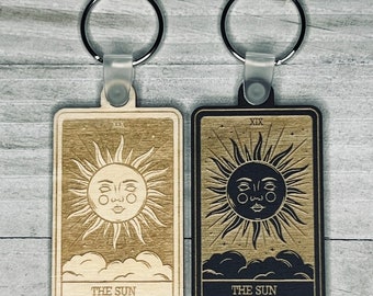 May include: Two wooden keychains featuring the The Sun tarot card. One keychain is light wood with a gold sun and the other is dark wood with a black sun.