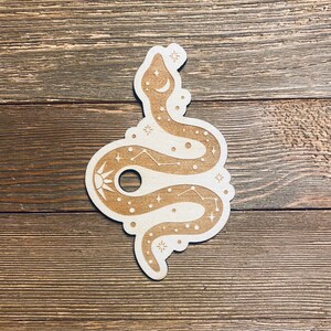 May include: A wooden cutout of a snake with a celestial design. The snake is curled up with its tail in its mouth, and the design includes stars, moons, and a sun.