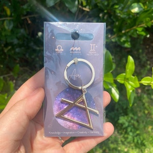 May include: Gold keychain with a triangle symbol and the word "Air" on a ring. The triangle is on a background with a galaxy design. The card behind the keychain lists the zodiac signs Libra, Aquarius, and Gemini with their dates.