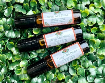 May include: Three brown glass roll-on bottles with black caps, labeled "Sleep Well Blend." The labels include text about ingredients like Clary Sage and Chamomile. The bottles are arranged on a bed of green leaves.