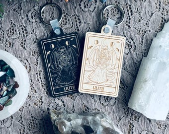 May include: Two rectangular keychains with metal rings. One is black, the other is natural wood. Both feature an engraved illustration of a figure with wings, a crescent moon, and the word "LILITH".
