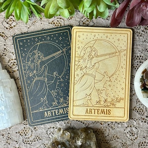 May include: Two wooden tarot cards with a black and white design of a woman with a bow and arrow, surrounded by deer. The cards both say "ARTEMIS" at the bottom.