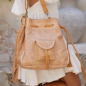 May include: A tan leather bucket bag with intricate embossed patterns and a drawstring closure. The bag has a long strap and a front pocket with a tassel.