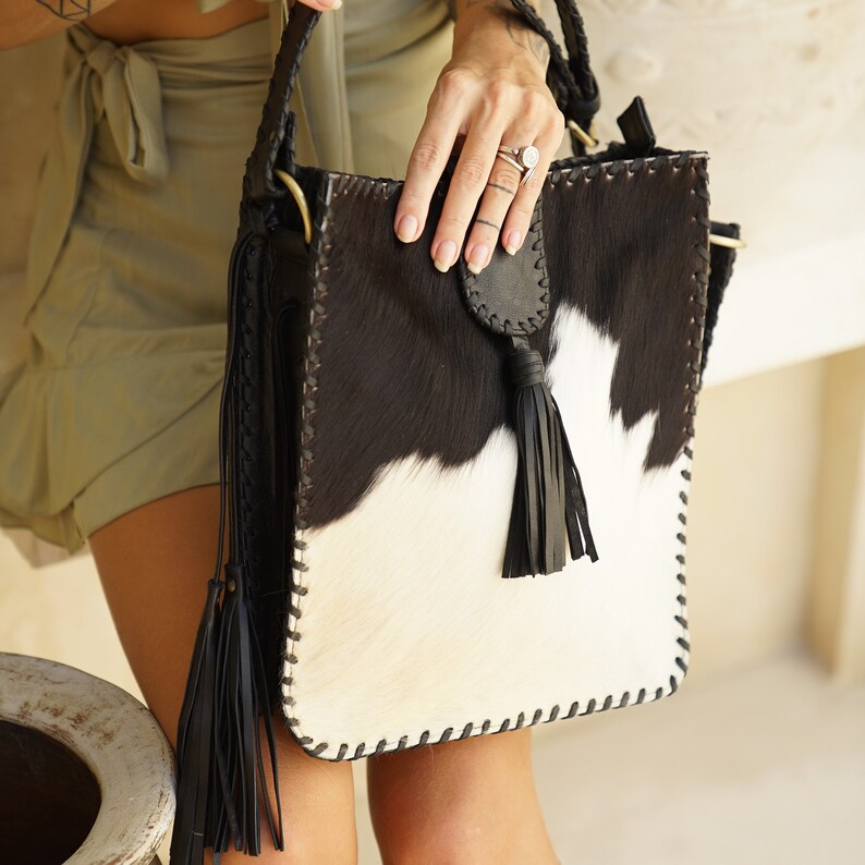 May include: A black and white cowhide tote bag with a black tassel. The bag has a stitched design around the edges and a black leather strap.