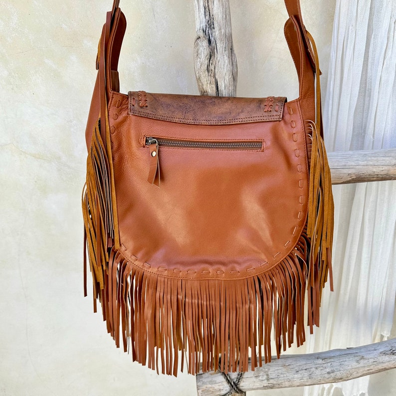 May include: A brown leather shoulder bag with fringe detailing. The bag has a zippered pocket on the front and a long strap.