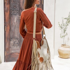 May include: A rust-coloured, long-sleeved top and tiered maxi skirt ensemble. A fringed, cowhide leather shoulder bag with brown and white patches hangs from the shoulder. The bag has a cream-coloured strap and fringe.