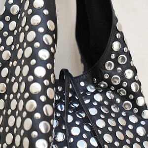 May include: Black leather shoulder bag with numerous silver-colored, circular studs. The bag has a soft, slouchy design and a long strap. The studs are evenly spaced across the surface, creating a bold, textured look.