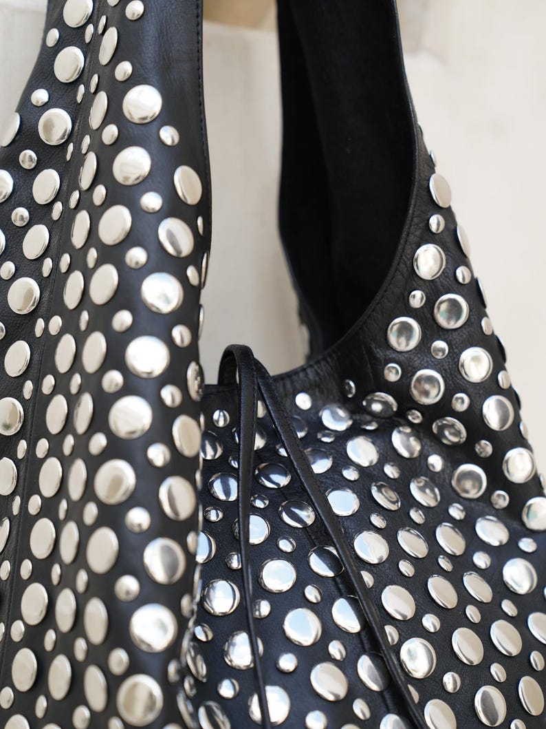 May include: Black leather shoulder bag with numerous silver-colored, circular studs. The bag has a soft, slouchy design and a long strap. The studs are evenly spaced across the surface, creating a bold, textured look.