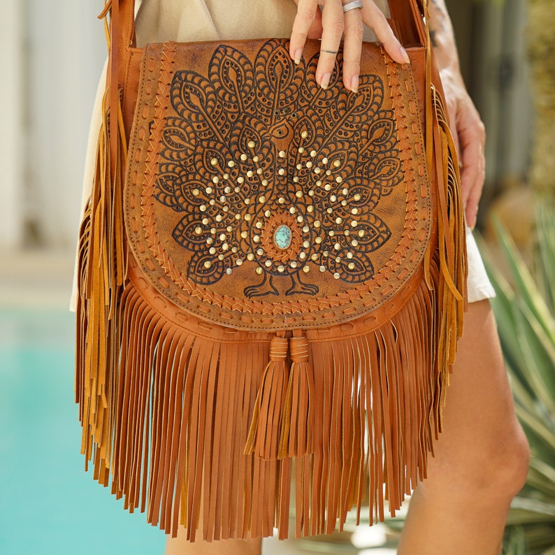 May include: A brown leather crossbody bag with a peacock design and turquoise stone accents. The bag has a long fringe and a tassel.