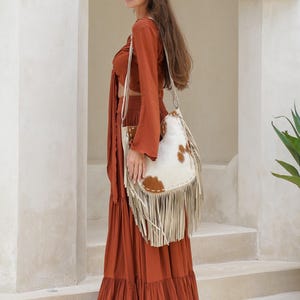 May include: A rust-coloured two-piece outfit with a long, tiered skirt and a long-sleeved crop top. A white and brown cowhide shoulder bag with fringe hangs from the shoulder. The bag has a cream-coloured strap.