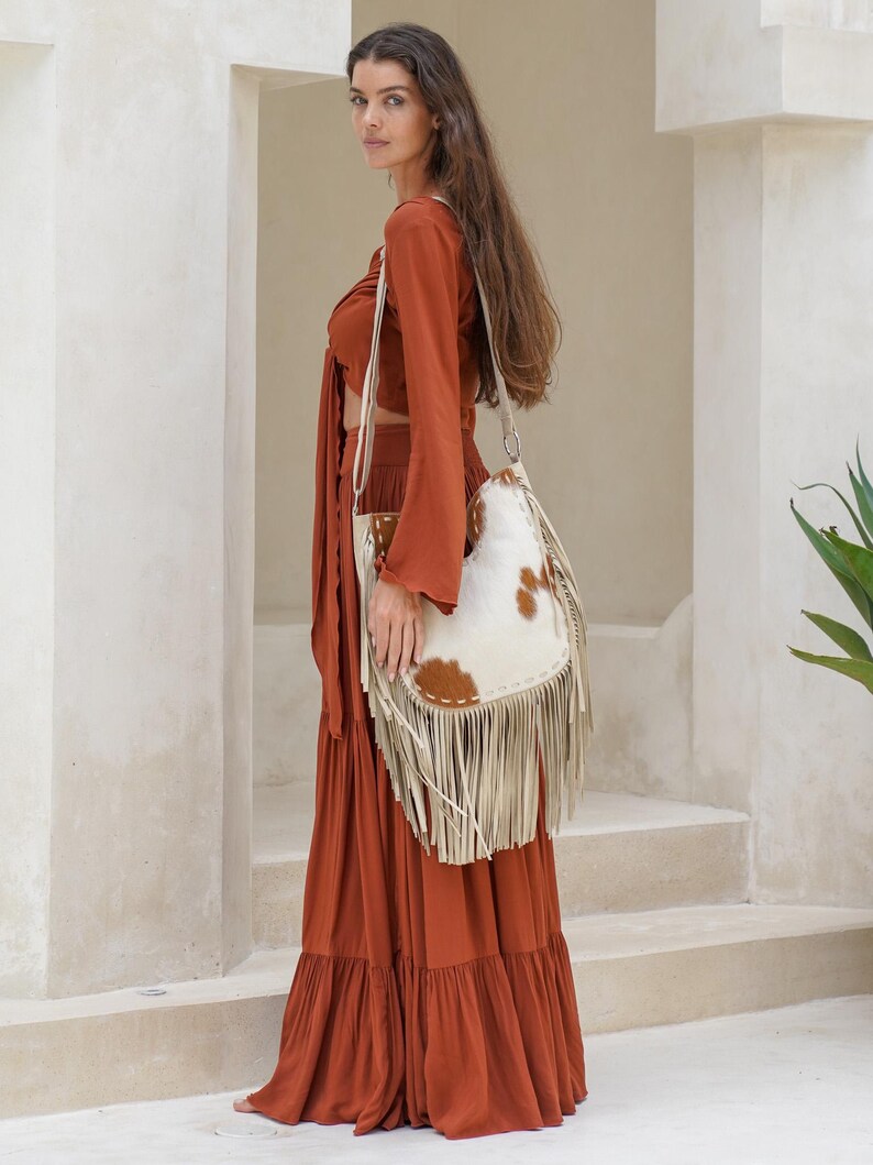 May include: A rust-coloured two-piece outfit with a long, tiered skirt and a long-sleeved crop top. A white and brown cowhide shoulder bag with fringe hangs from the shoulder. The bag has a cream-coloured strap.