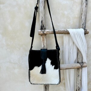 May include: Black and white cowhide crossbody bag with a black leather tassel. The bag has a stitched edge and is hanging on a wooden ladder.