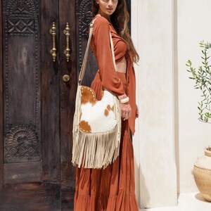 May include: A rust-coloured outfit with a long skirt and a matching top. A white and brown cowhide shoulder bag with fringe hangs from the shoulder. The background includes a wooden door and a white wall.