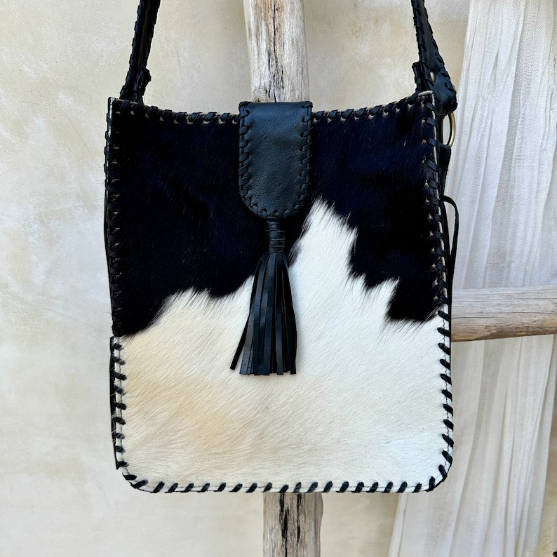 May include: Black and white cowhide crossbody bag with a black leather tassel. The bag has a stitched edge and a black strap.