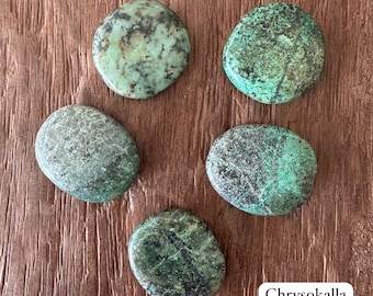 以下が含まれることがあります： 緑とターコイズの色合いの、磨かれたクリソコラ石が5つ。濃い茶色と黒の縞模様があります。石は滑らかで丸みを帯びており、自然で素朴な外観です。「Chrysokalla」の文字が右下の白いボックスにあります。