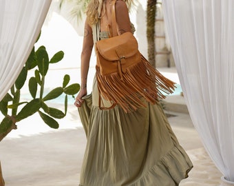 May include: A tan leather backpack with a flap closure and fringe detail. The bag is worn over a long, olive green dress. The image is taken outdoors with natural light, and the background includes white curtains and greenery.