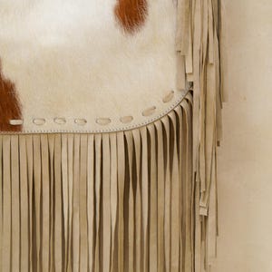 May include: Close-up of a cowhide bag with a cream body and brown spots. The bag has a cream fringe along the bottom edge and a stitched seam detail. The bag is a bohemian style.