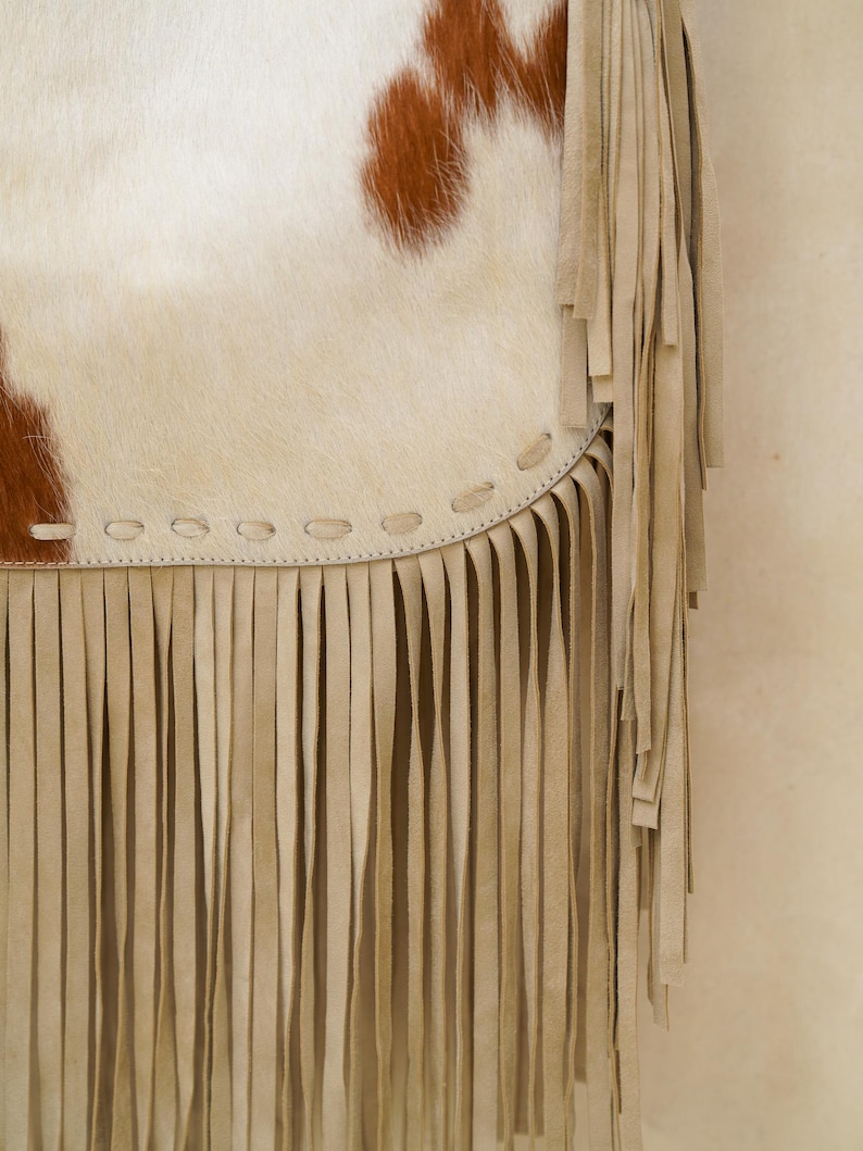 May include: Close-up of a cowhide bag with a cream body and brown spots. The bag has a cream fringe along the bottom edge and a stitched seam detail. The bag is a bohemian style.