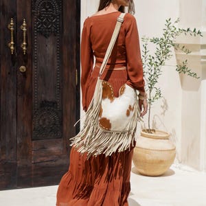 May include: A rust-coloured long-sleeve top and tiered maxi skirt ensemble. A cream-coloured shoulder bag with a cowhide front and fringe detailing is worn over the shoulder. The outfit is worn outdoors.