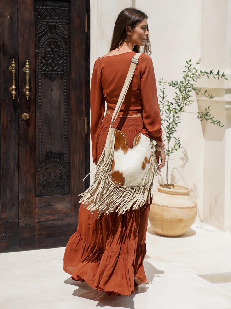 May include: A rust-coloured long-sleeve top and tiered maxi skirt ensemble. A cream-coloured shoulder bag with a cowhide front and fringe detailing is worn over the shoulder. The outfit is worn outdoors.