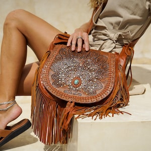 May include: A brown leather satchel bag with a peacock design and fringe. The bag has a turquoise stone embellishment and is decorated with metal studs.