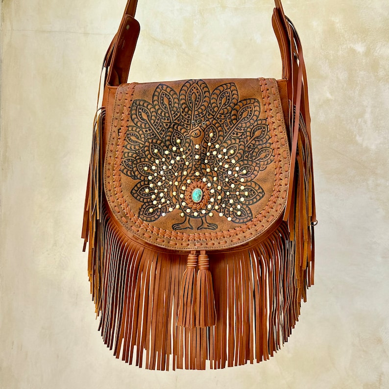 May include: A brown leather shoulder bag with a peacock design and fringe. The peacock is embellished with rhinestones and a turquoise stone.