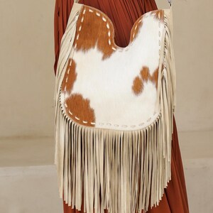 May include: A cream leather shoulder bag with a cowhide front panel. The cowhide is brown and white. The bag has long cream fringe along the bottom and a cream strap.