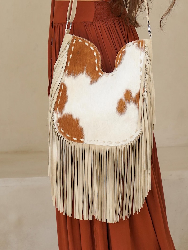 May include: A cream leather shoulder bag with a cowhide front panel. The cowhide is brown and white. The bag has long cream fringe along the bottom and a cream strap.