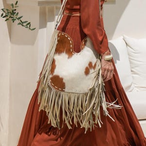 May include: A shoulder bag with a cowhide pattern in brown and white, featuring cream-coloured fringe and a strap. The bag is carried over the shoulder. The background is a white wall and a person wearing a rust-coloured dress.