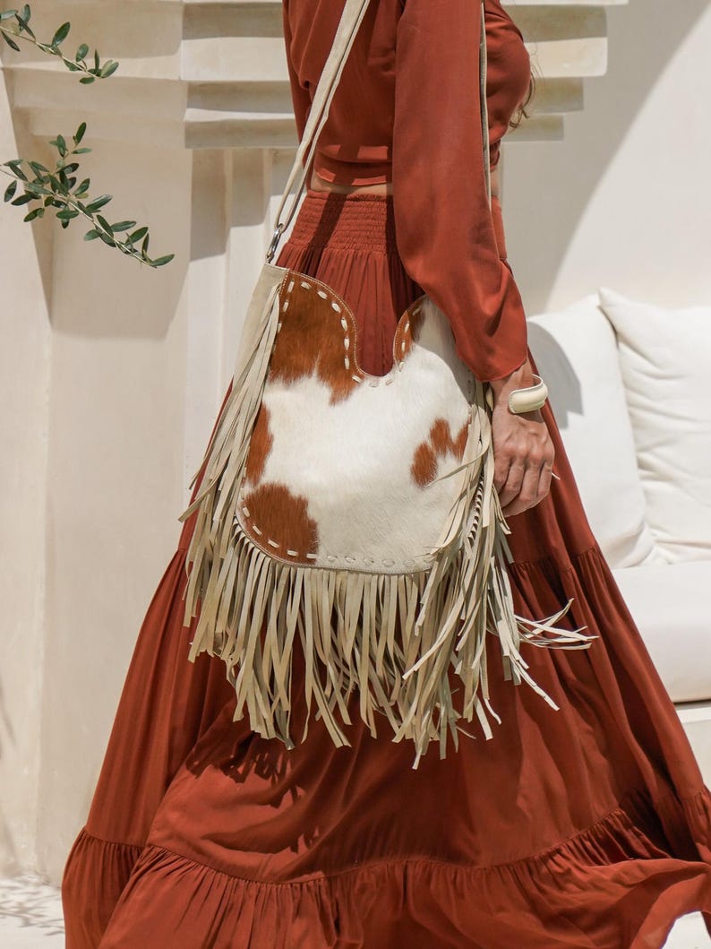 May include: A shoulder bag with a cowhide pattern in brown and white, featuring cream-coloured fringe and a strap. The bag is carried over the shoulder. The background is a white wall and a person wearing a rust-coloured dress.