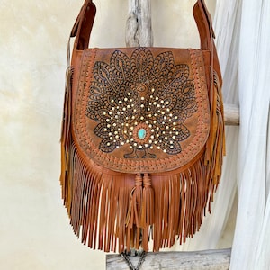 May include: Brown leather shoulder bag with a peacock design and fringe. The bag has a turquoise stone accent.