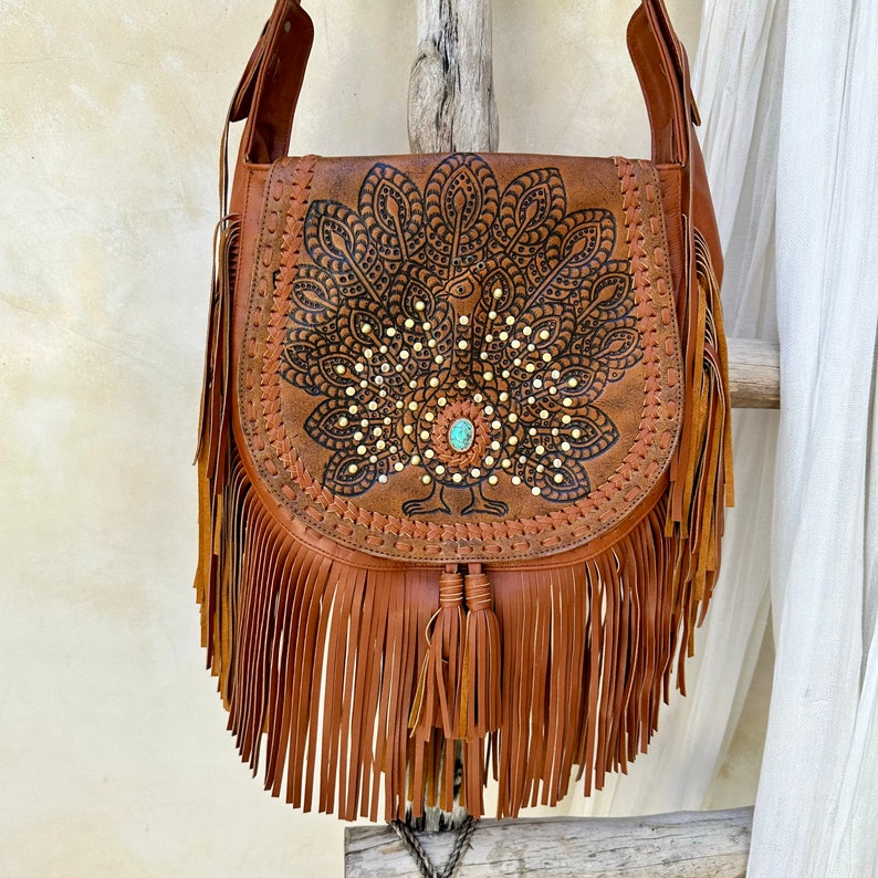 May include: Brown leather shoulder bag with a peacock design and fringe. The bag has a turquoise stone accent.