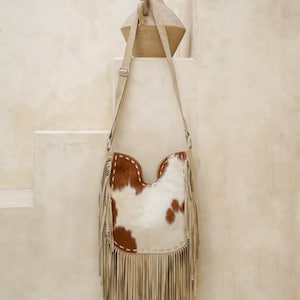 May include: A cowhide leather shoulder bag with a cream-coloured fringe. The bag features a white and brown spotted pattern and a cream-coloured adjustable strap. The bag is hanging on a wall.