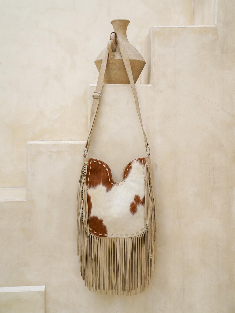 May include: A cowhide leather shoulder bag with a cream-coloured fringe. The bag features a white and brown spotted pattern and a cream-coloured adjustable strap. The bag is hanging on a wall.