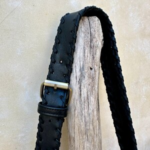 May include: Black leather strap with a braided design and a gold metal buckle. The strap is hanging on a piece of driftwood.