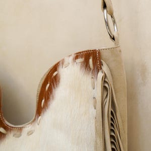 May include: A cream-coloured shoulder bag with a cowhide pattern and fringe detailing. The bag features a brown leather trim with white stitching and a silver metal ring. The bag's design is a mix of textures and colours.