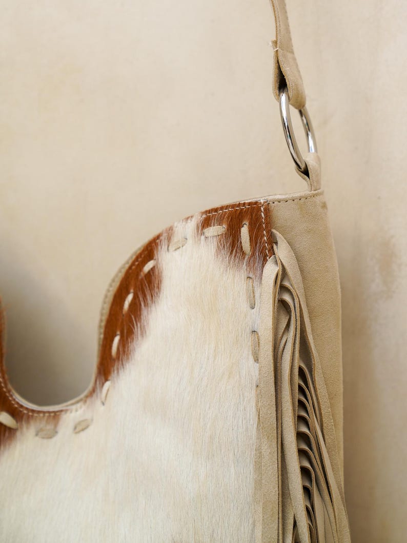 May include: A cream-coloured shoulder bag with a cowhide pattern and fringe detailing. The bag features a brown leather trim with white stitching and a silver metal ring. The bag's design is a mix of textures and colours.