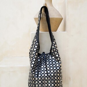 May include: Black leather shoulder bag adorned with numerous silver-colored studs. The bag has a drawstring closure and a single shoulder strap. The bag is hanging against a neutral-colored background.