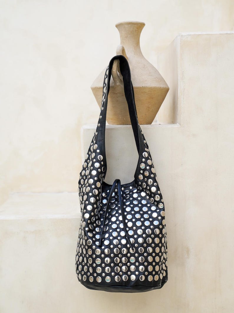 May include: Black leather shoulder bag adorned with numerous silver-colored studs. The bag has a drawstring closure and a single shoulder strap. The bag is hanging against a neutral-colored background.