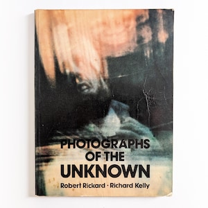 May include: A vintage book titled "Photographs of the Unknown" by Robert Rickard and Richard Kelly. The cover displays a blurred, abstract image with a dark backdrop and the title in bold, white letters. The book shows signs of wear.